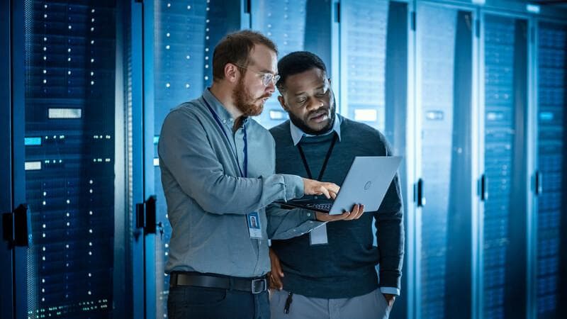 Bearded IT Technician in Glasses with Laptop Computer and Black Male Engineer Colleague are Using Laptop in Data Center while Working Next to Server Racks. Running Diagnostics or Doing Maintenance Work