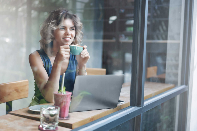 Attractive stylish female entrepreneur working outside office sit cafe look outside window passersby dreamy smiling holding cup coffee enjoy momentum working laptop prepare project